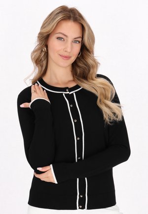 Młoda kobieta z długimi falującymi blond włosami, ubrana w czarny kardigan z białym obszyciem i guzikami, pozująca z rękami skrzyżowanymi na białym tle.