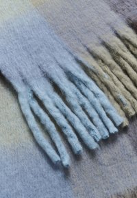 Uldhalstørklæde med lange, slanke lyseblå frynser mod en gradient baggrund af lyseblå og beige nuancer. Blødt, tekstureret stof.