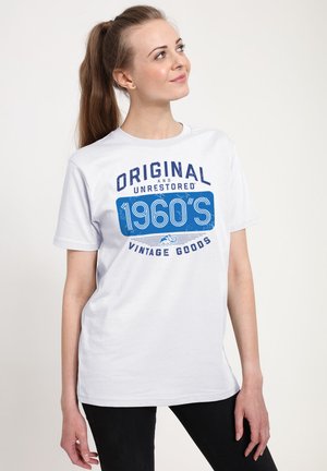 Junge Frau mit langem Pferdeschwanz, die ein weißes T-Shirt mit blauem Text "Original und unrestaurierte Vintage-Waren aus den 1960er Jahren" trägt, blickt nach rechts oben.