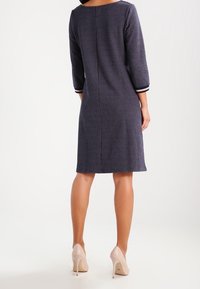 Marinblå klänning med strukturerat mönster, trekvartsärmar och kontrasterande ränder vid manschetterna, knälång längd, bärs med nakna klackar.