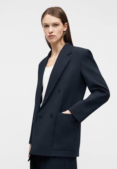 Γυναίκα με ίσια καστανά μαλλιά φοράει σκούρο διπλόκουμπο σακάκι πάνω από λευκό τοπ, με το ένα χέρι στην μεγάλη μπροστινή τσέπη του σακακιού.