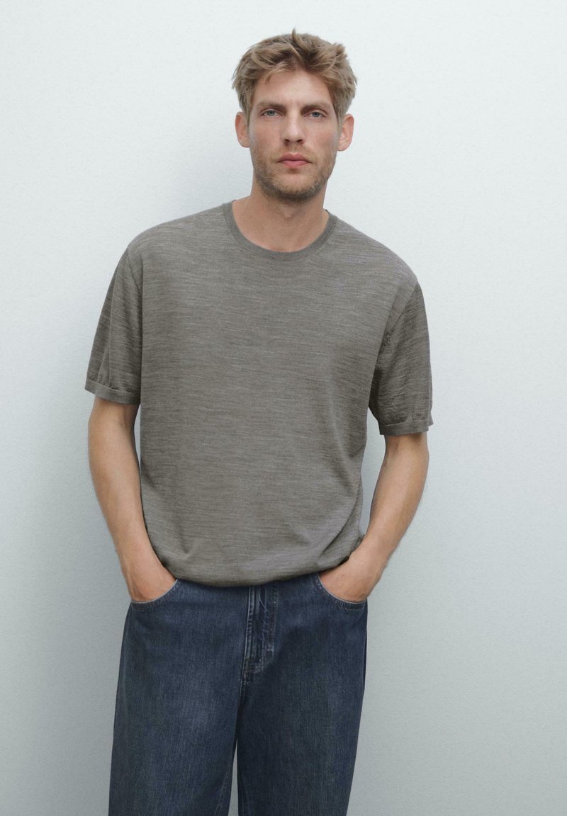 Grå kortærmet t-shirt med rund hals, lavet af tekstureret stof, parret med blå denim jeans. Hænderne afslappet i lommerne.