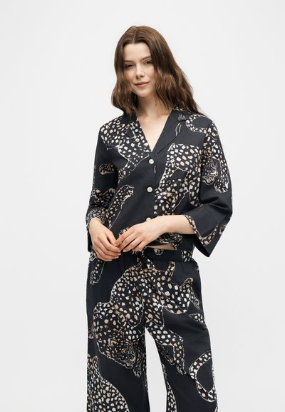Γυναίκα με κυματιστά καστανά μαλλιά που φοράει μαύρο σετ πιτζάμες με μπεζ και λευκό λεοπάρ μοτίβο, στέκεται μπροστά σε απλό ανοιχτό φόντο.