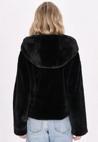 Giacca nera in pelliccia sintetica con cappuccio, maniche lunghe, vestibilità comoda e texture morbida, abbinata a jeans azzurri chiari.