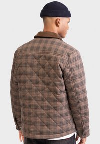 Mand iført en brun quiltet plaidjakke og en sort strikkebehætte, vist fra baggrunden mod en ensfarvet lys baggrund.