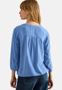 Blouse bleu clair, ample, avec des manches 3/4, en tissu texturé et un décolleté arrondi, avec une pince dans le dos pour un meilleur tombé.