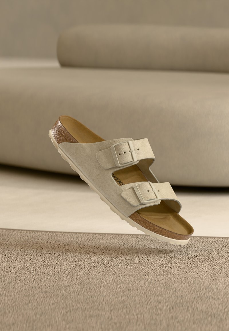 Μπεζ σανδάλι από σουέντ με δύο ρυθμιζόμενες αγκράφες και φελό πάτο, αιωρούμενο πάνω από υφή μπεζ χαλιού και απαλού μπεζ επίπλου.