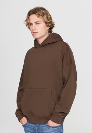 Felpa marrone con tasca a canguro, grande cappuccio e polsini a costine. Il tessuto ha una texture liscia e un fit rilassato. Indossata con jeans blu.