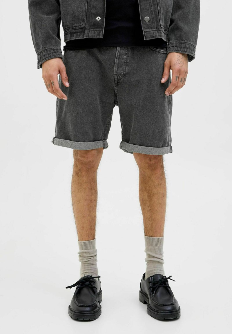 Shorts en denim noir avec revers, associés à des chaussettes gris clair et des chaussures à lacets noires. Les shorts ont une coupe décontractée et une texture subtile.