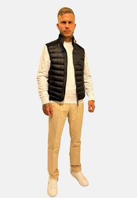 Gilet rembourré noir, chemise à manches longues blanche, pantalon beige et baskets blanches. Le gilet a une fermeture zippée à l'avant et des poches latérales.