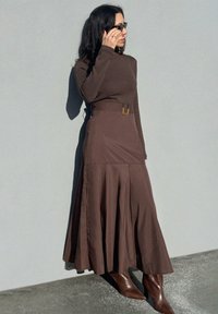 Robe longue marron à manches longues avec une jupe évasée, dotée d'un détail de boucle à la taille. Un tissu lisse et des bottes marron jusqu'aux genoux complètent le look.