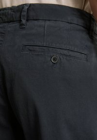 Pantalons bleu marine foncé avec une texture lisse, passants de ceinture, une poche arrière avec un bouton et un détail logo subtil sur le tissu.