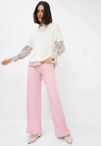 Haut blanc avec des manches rayées superposé sur un pantalon large rose. Les chaussures sont de couleur beige clair avec un bout pointu. Texture de tissu lisse.
