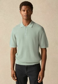 Light green knit polo shirt with short sleeves, textured pattern, and a zipper at the collar. Worn with dark jeans.