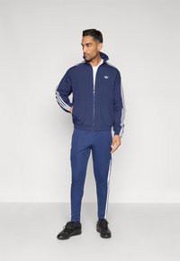 Námornícka tepláková súprava s bielymi pruhmi na rukávoch a nohaviciach, vyrobená z hladkej látky; bunda na zips a priliehavé nohavice, doplnené čiernymi teniskami.