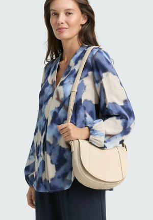 Borsa a tracolla in pelle beige con pattina curva, tracolla regolabile e superficie texture; indossata con una blusa a fantasia astratta blu e bianca.