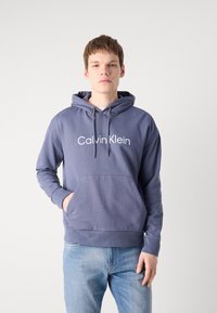 Felpa leggero blu-grigiastro con coulisse e grande tasca frontale, caratterizzata dal logo bianco "Calvin Klein" in un font moderno. Tessuto morbido.