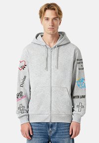 Halli tõmblukuga kapuutsiga pusa esitaskutega, mille varrukatel on mitmevärvilised graafilised mustrid ja tekstid, sealhulgas südamed ja ristiembleem.