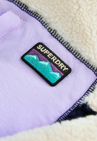 Ljubičasta tkanina s teksturiranim kremastim podstavljanjem ima crni aplikator s natpisom "SUPERDRY" te dizajn planina u tirkiznoj i ljubičastoj boji.