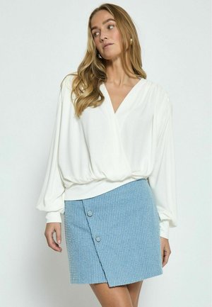 Blusa bianca a maniche lunghe con scollatura a V e fondo arricciato, abbinata a una mini gonna blu con texture e due bottoni laterali.