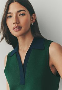 Pull polo sans manches vert avec un col marine ; matière en tricot texturée ; design en V ; modèle portant des boucles d'oreilles en argent.