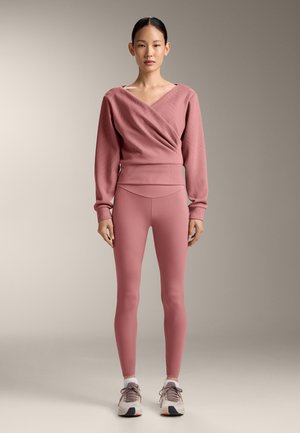 Vrouw staat naar voren gericht en draagt een langärmelig overslagtop in stofroze, bijpassende legging, witte sokken en beige sneakers.