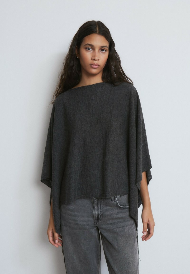 Poncho suelto de color gris oscuro, con escote amplio, tela texturizada y mangas cortas. Combinado con jeans de pierna recta en gris claro.