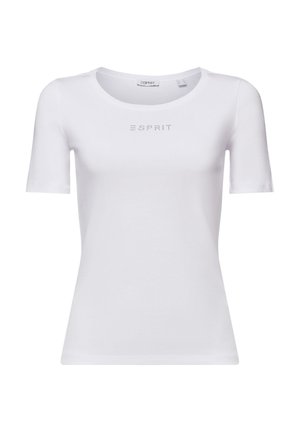 Femme portant un haut blanc à manches courtes avec texte, pantalon plissé gris et bijoux en or, tenant une sangle métallique de sac à épaule.