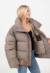 Giacca puffer color taupe con colletto alto, dotata di trapuntatura orizzontale, zip frontale e una toppa arancione sul lato sinistro.