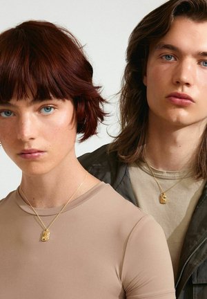 Goldene Halsketten mit rechteckigen Anhängern an Personen, die enganliegende beige und graue Oberteile tragen, vor einem schlichten Hintergrund.