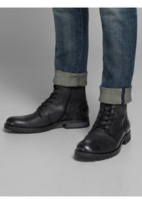 Zwarte leren laarzen gedragen met opgerolde denim jeans, wat een casual en robuuste stijl benadrukt. De achtergrond is neutraal grijs.