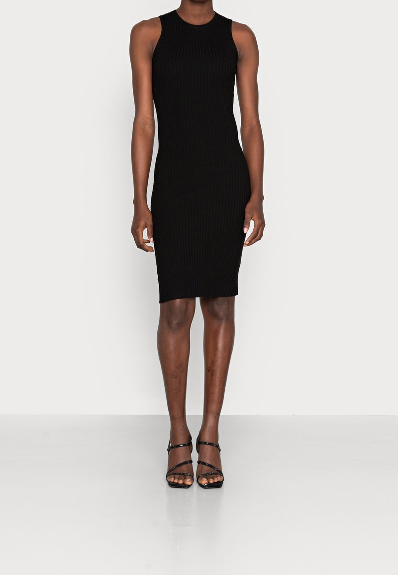 Femme portant une robe noire moulante sans manches à côtes, associée à des sandales noires à talons hauts et à brides, debout sur un fond clair uni.