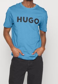 Niebieska bawełniana koszulka z wyrazistym czarnym logo "HUGO" z przodu, okrągłym dekoltem oraz krótkimi rękawami, w zestawie z czarnymi spodniami.