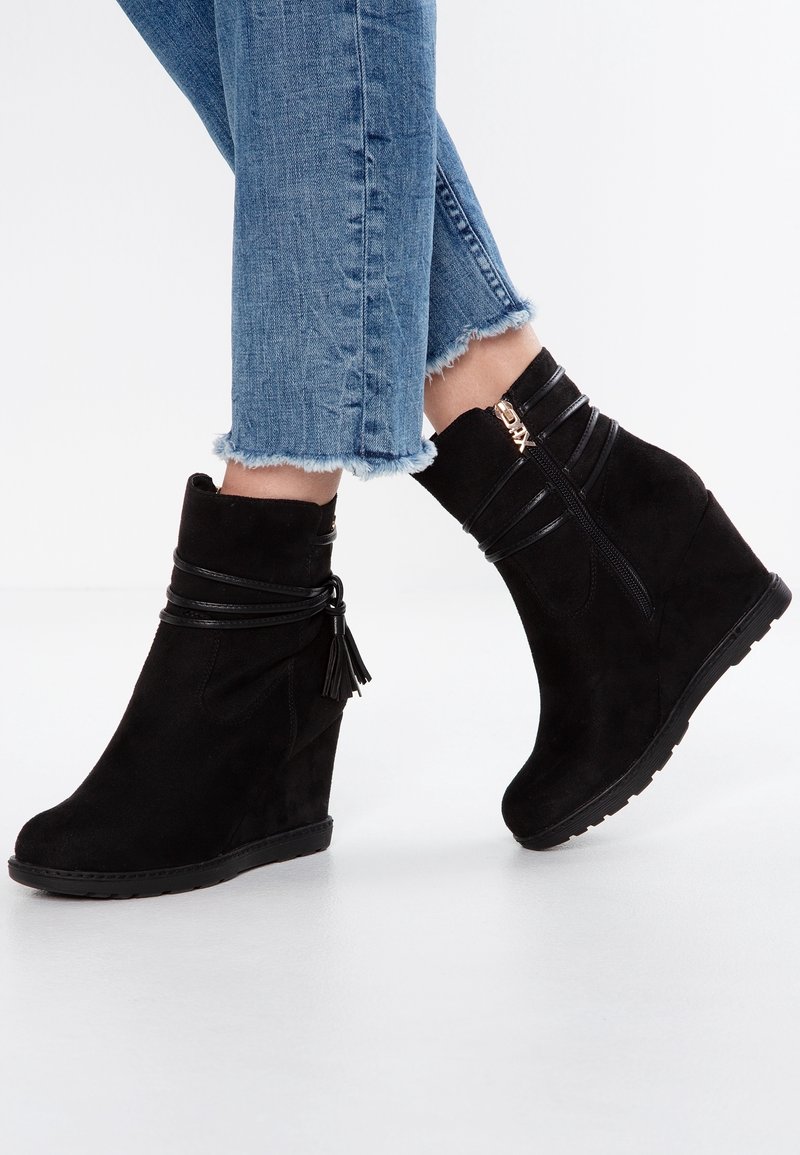 Stivali alla caviglia neri in suede con tacco a cuneo presentano una nappina decorativa, cinturini in pelle e una zip laterale. Abbinati a jeans in denim con orlo sfrangiato.