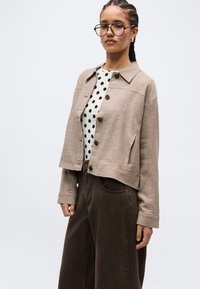 Ženska, oblečena v bež jakno z gumbi, belo srajco s črnimi pikami, temne široke hlače, okrogle očala in spletene lase na belem ozadju.