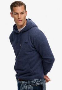 Sudadera con capucha azul marino en suave forro polar con un bolsillo frontal y cordón ajustable. Cuenta con un pequeño logo en el pecho y un ajuste relajado. Se lleva con jeans.
