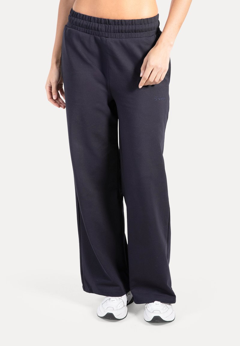 Marineblauwe wijduitlopende joggingbroek met elastische tailleband en zijzakken, gemaakt van zachte stof. Witte sneakers maken de look compleet.