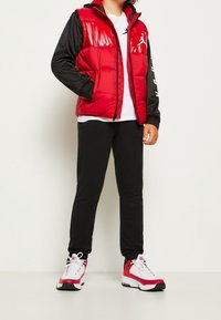 Rode puffer vest met glanzende afwerking, zwarte mouwen, witte T-shirt, zwarte trainingsbroek en rood/witte sneakers met zwarte accenten.