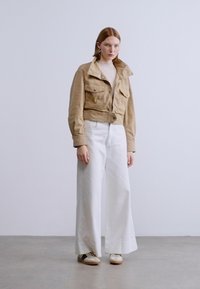 Σύντομο μπεζ σακάκι με δύο τσέπες στο στήθος και κούμπωμα με κουμπιά, συνδυασμένο με λευκό τζιν φαρδιάς γραμμής και μπεζ αθλητικά παπούτσια, τοποθετημένο σε απλό φόντο.