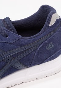 Gros plan d'une basket en daim bleu marine avec des bandes en cuir noir, marquage « GEL » sur le côté et semelle blanche amortissante.
