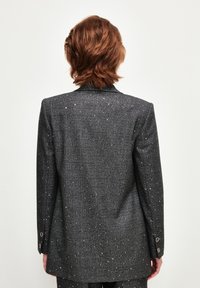 Blazer grigio con un motivo a quadri testurizzato, arricchito da piccole macchie metalliche e dettagli con bottoni sulle maniche. Vista posteriore.