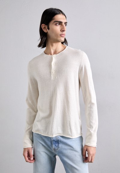 Jeune homme aux longs cheveux noirs portant une chemise henley à manches longues de couleur crème et un jean bleu clair, regardant sur le côté.