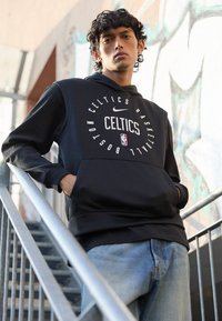 Schwarzer Hoodie mit "Celtics" und Basketballgrafiken, ausgestattet mit einer Kängurutasche. Der Modell steht auf Treppen und trägt dazu hellblaue Jeans.