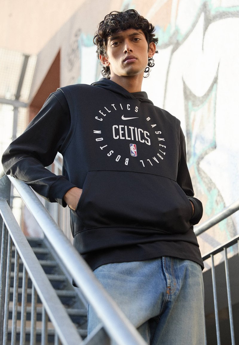 Schwarzer Hoodie mit "Celtics" und Basketballgrafiken, ausgestattet mit einer Kängurutasche. Der Modell steht auf Treppen und trägt dazu hellblaue Jeans.