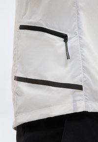 Section de veste blanche avec une poche zippée noire horizontale et une bande réfléchissante noire au-dessus de l'ourlet blanc, portée sur un pantalon noir.