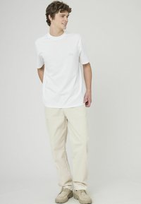 Jeune homme portant un t-shirt blanc à manches courtes, un pantalon beige clair et des chaussures beige, se tenant devant un fond clair uni.