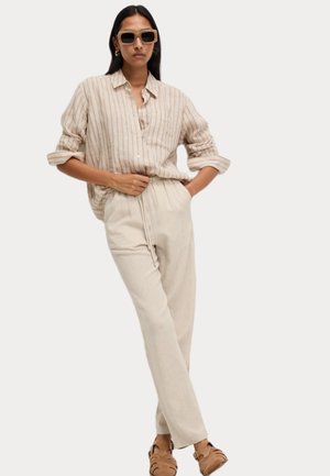Beige gestreiftes Hemd mit entspannter Passform, getragen mit hellbeigen hochgeschnittenen Hosen. Braune Riemchensandalen und quadratische Sonnenbrille.