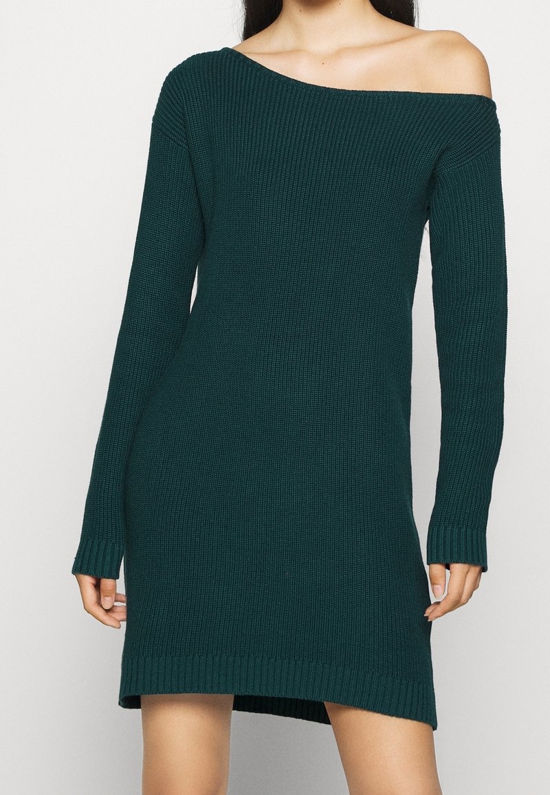 Abito a maglia a coste in un profondo color teal, con scollatura off-shoulder e maniche lunghe. L'orlo è leggermente più lungo sul retro.