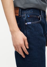 Hånd som hviler nær lommen på mørkeblå denimjeans med Red Bull Racing-merke og brun lærlapp på midjebåndet.