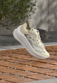 Μπεζ και λευκό αθλητικό παπούτσι Calvin Klein που αιωρείται πάνω από ξύλινο πλακάκι, με πράσινα φύλλα και γκρι τοίχο στο φόντο.